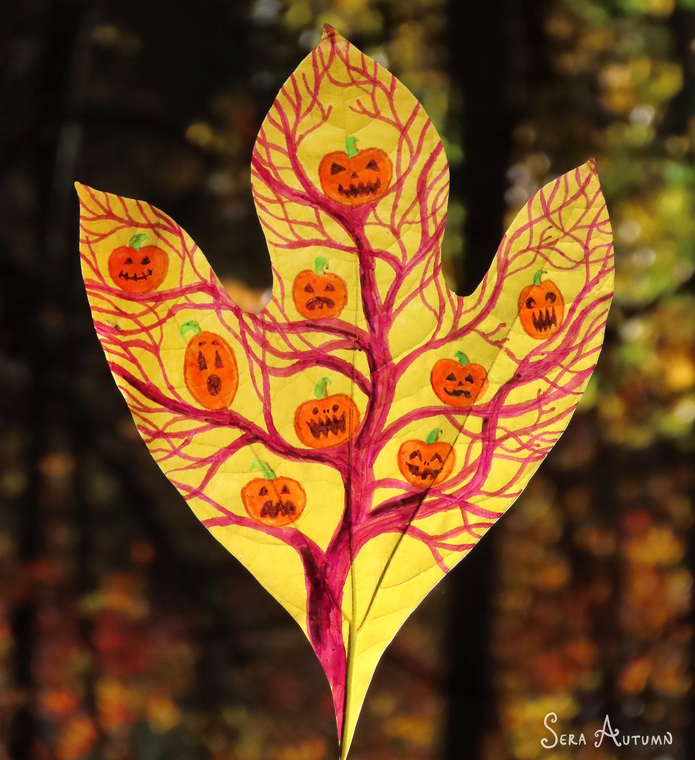 A bare pink tree hung with grinning jack-o'-lanterns, drawn on a yellow sassafras leaf against autumn bokeh, by Sera Autumn