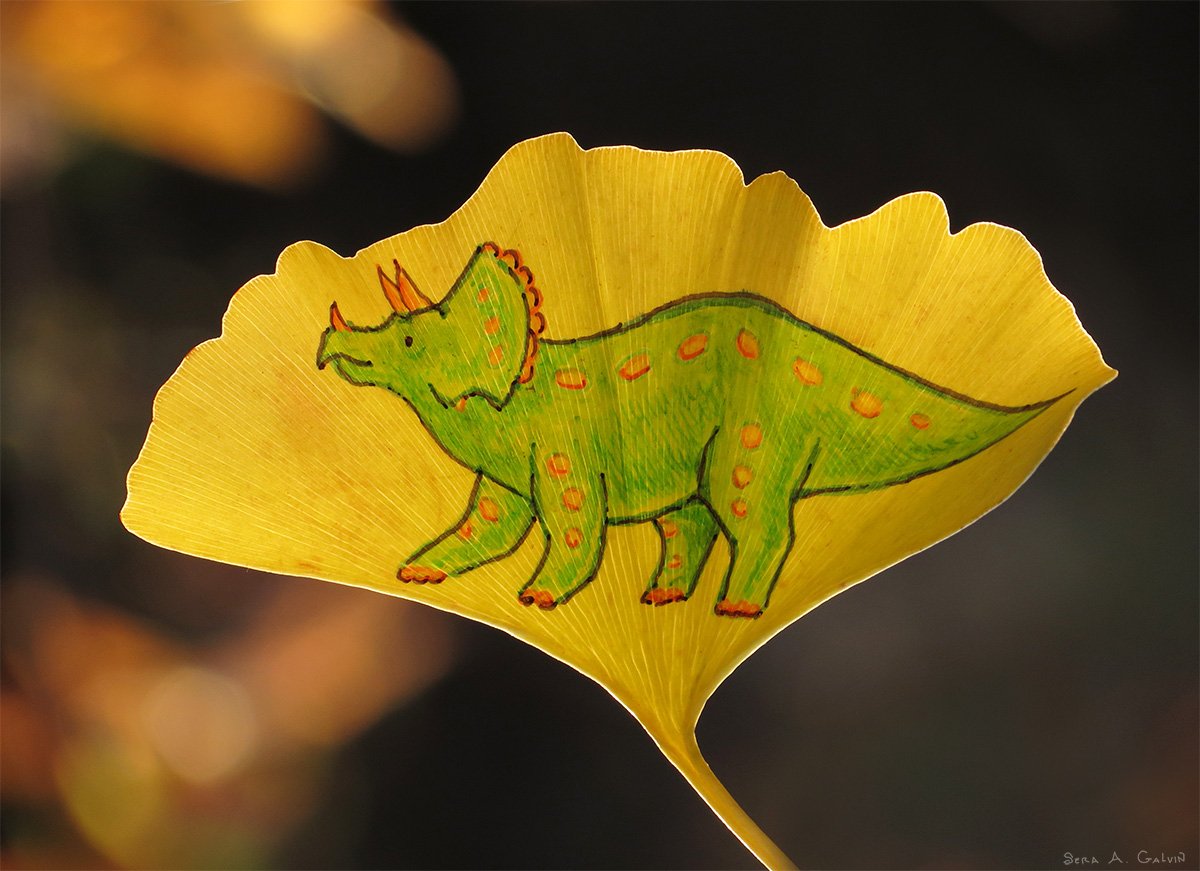 A green orange-spotted triceratops dinosaur drawn on a yellow ginkgo leaf against a dark blurred background, by Sera Autumn