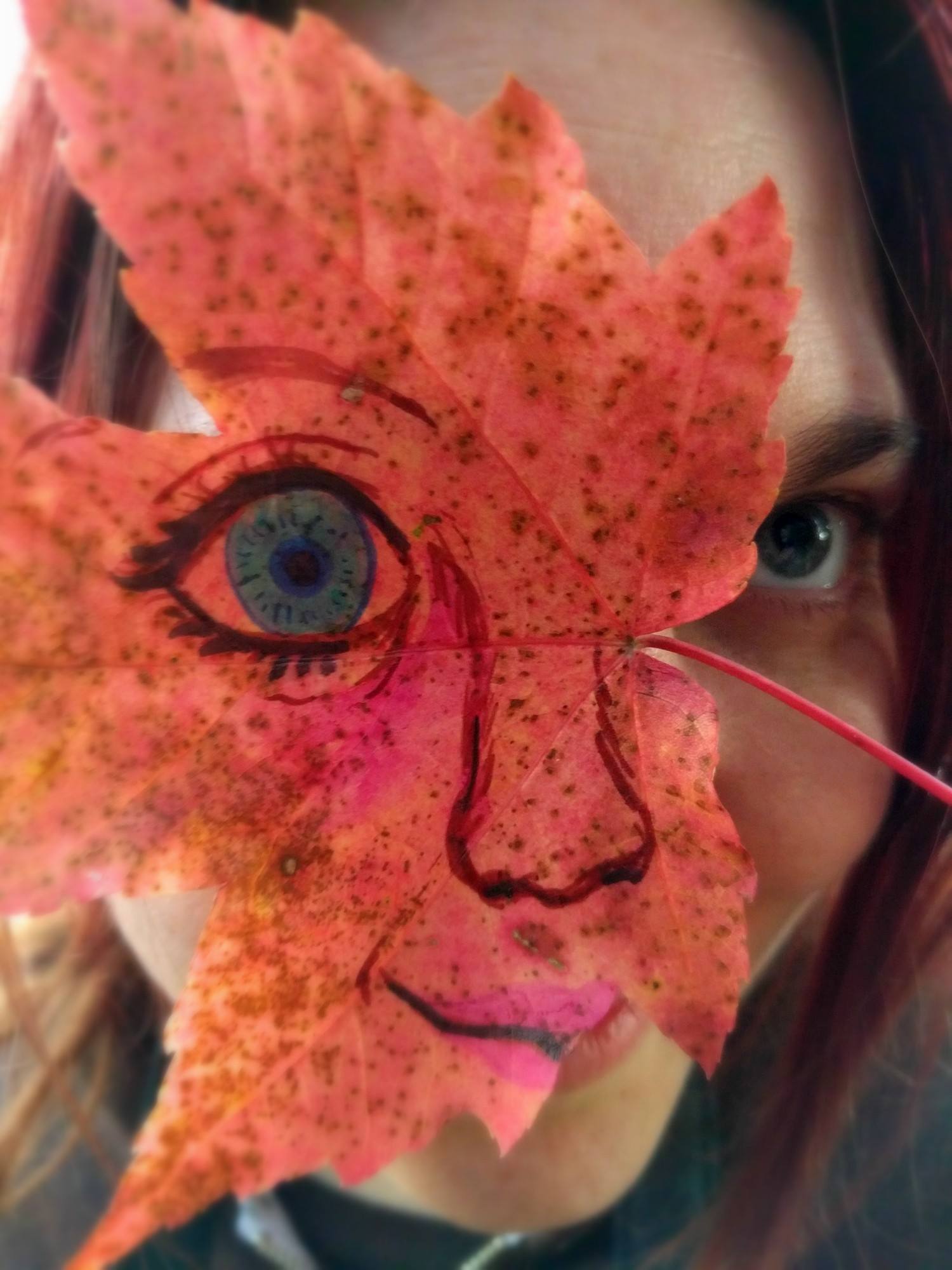 A large blue eye and face drawn on a red maple leaf held over a mysterious woman's real face, by Sera Autumn
