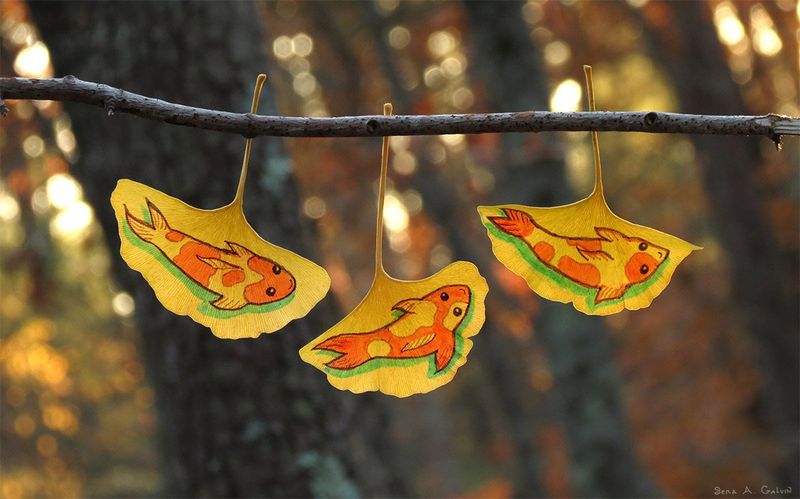 Three yellow ginkgo leaves drawn with orange koi fish, hung from a bare branch against golden forest bokeh, by Sera Autumn