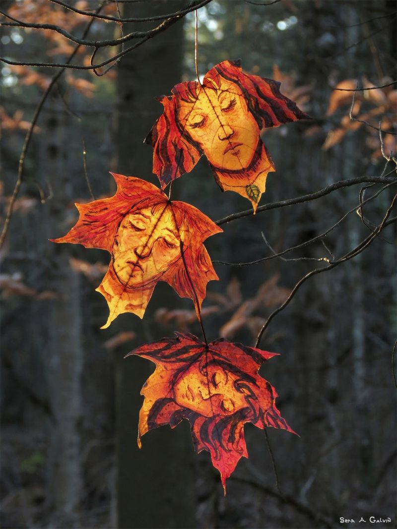 Three women's faces with closed eyes and flowing hair drawn on glowing orange-red maple leaves hanging in a dark forest, by Sera Autumn