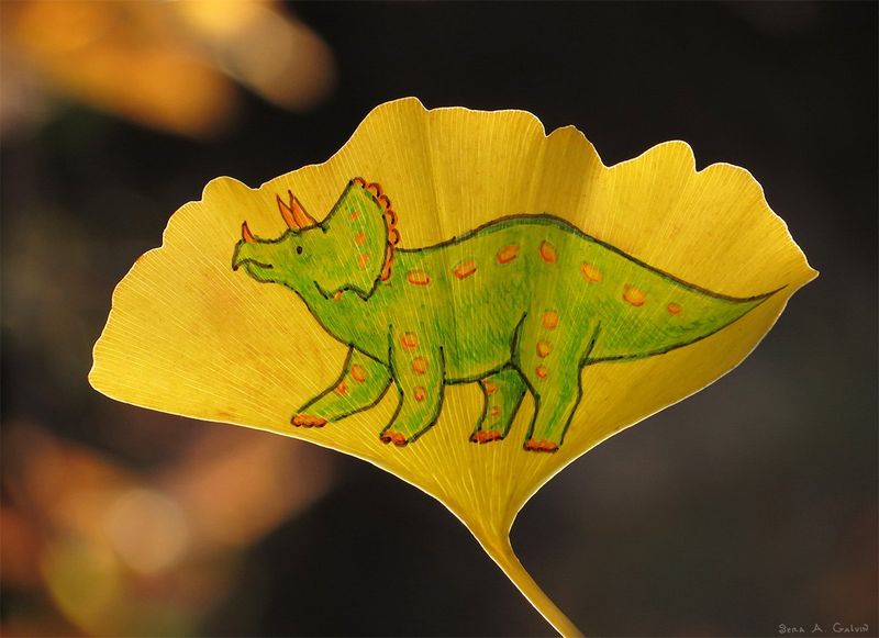 A green orange-spotted triceratops dinosaur drawn on a yellow ginkgo leaf against a dark blurred background, by Sera Autumn