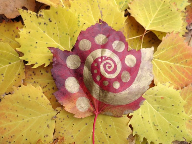 A gold spiral surrounded by pale dots drawn on a deep-red maple leaf resting atop a bed of yellow-green autumn leaves, by Sera Autumn