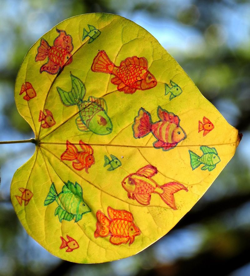 Many colorful goldfish in red, orange, and green swimming across a heart-shaped yellow leaf against blue-sky bokeh, by Sera Autumn