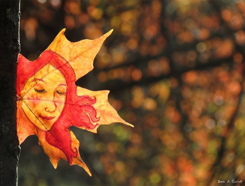 A serene woman's face with flowing red-gold hair drawn on a maple leaf against blurred autumn bokeh beside a tree trunk, by Sera Autumn