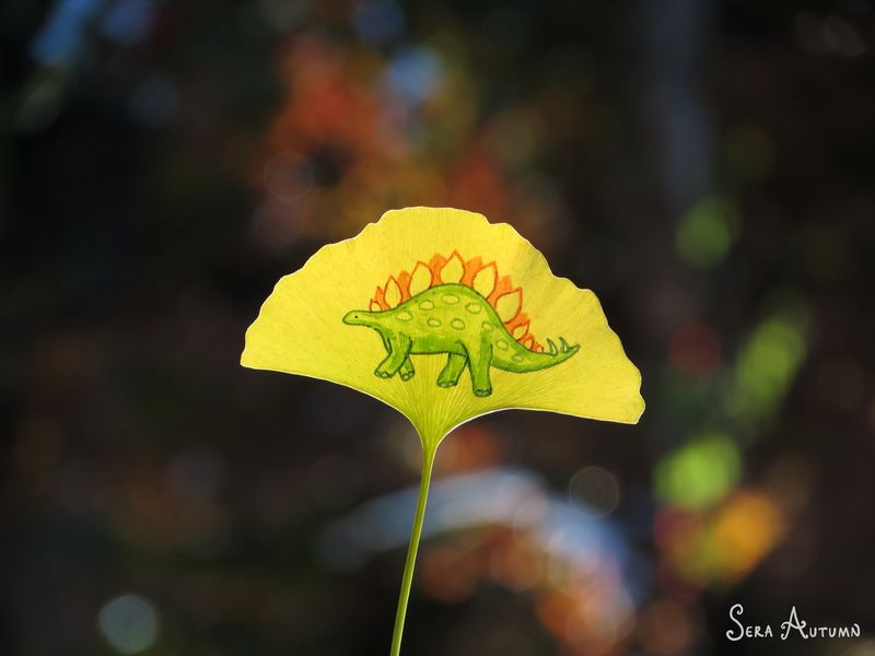 A green stegosaurus with orange back plates drawn on a bright yellow ginkgo leaf against dark bokeh, by Sera Autumn